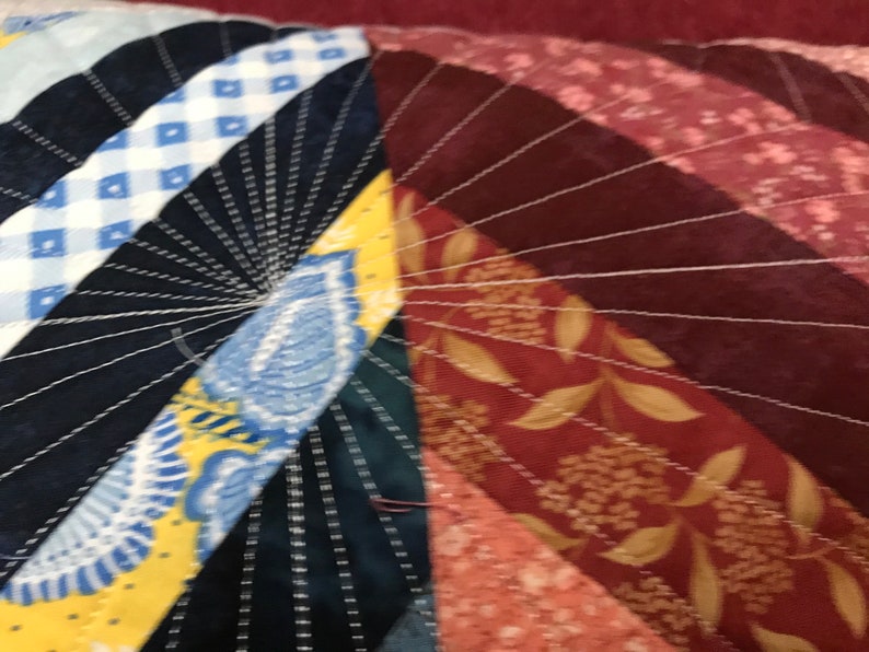 Puede incluir: Primer plano de una colcha con una variedad de telas y colores, incluyendo azul, amarillo, rojo y blanco. La colcha est&aacute; cosida en un patr&oacute;n radial, con las l&iacute;neas de costura que irradian desde el centro.