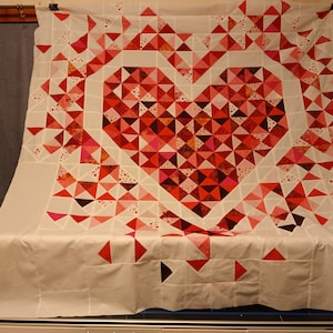 May include: A white quilt with a large red heart made of smaller red, pink, and white triangles. The heart is surrounded by a white border and the quilt is finished with a white backing.
