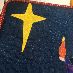 May include: A blue quilted fabric with a yellow star and a small orange and white candle. The fabric is quilted with a small, white, dotted pattern.