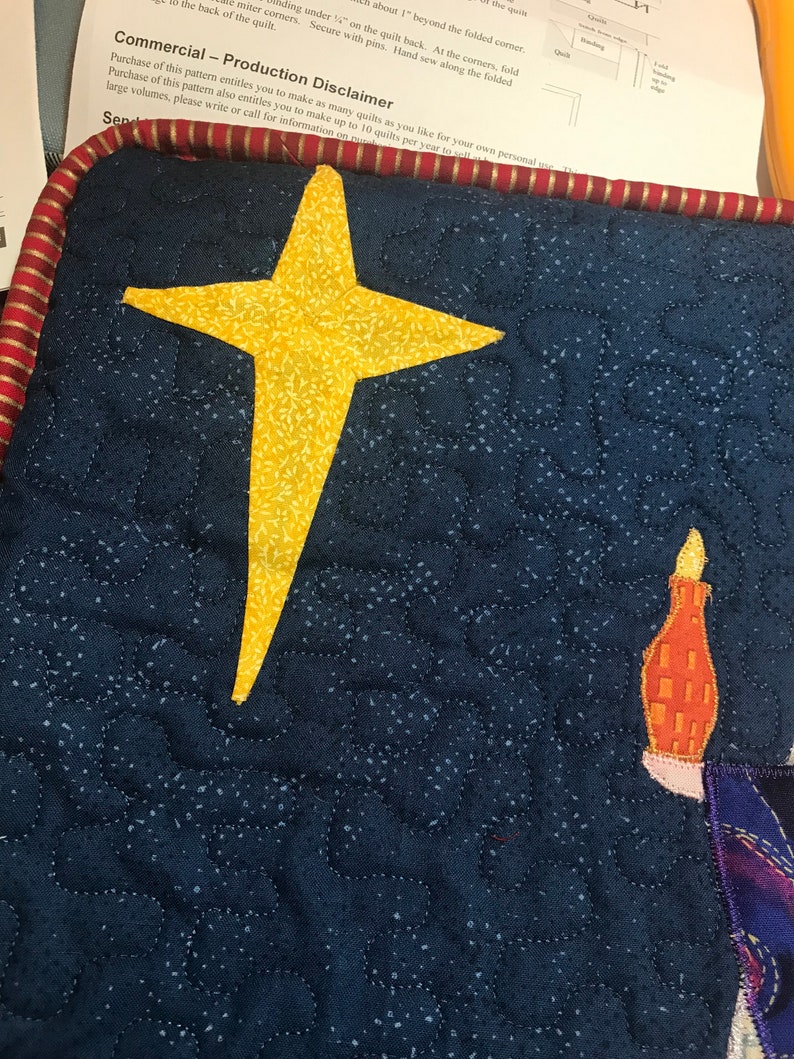 May include: A blue quilted fabric with a yellow star and a small orange and white candle. The fabric is quilted with a small, white, dotted pattern.
