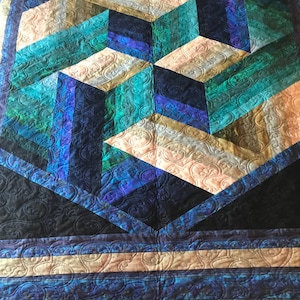 May include: A quilt with a geometric design featuring a 3D cube pattern in shades of blue, green, and beige. The quilt is quilted with a paisley pattern.