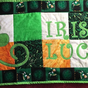 以下が含まれることがあります： 緑、白、オレンジのパッチワークのキルトで、「Irish Luck」という文字が緑色の刺繍で施されています。キルトの左上には大きな緑色のクローバーがあります。