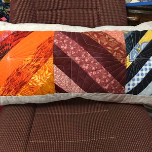 Puede incluir: Una almohada rectangular larga con una funda de tela gris. La almohada est&aacute; hecha de tela de patchwork en tonos naranja, rojo y azul. La tela est&aacute; cosida en un patr&oacute;n geom&eacute;trico.