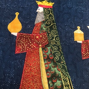 May include: A quilted Christmas stocking featuring a wise man holding a gold gift box. The wise man is wearing a red and gold robe with a green and gold patterned cape. The background is a dark blue fabric with a star pattern.