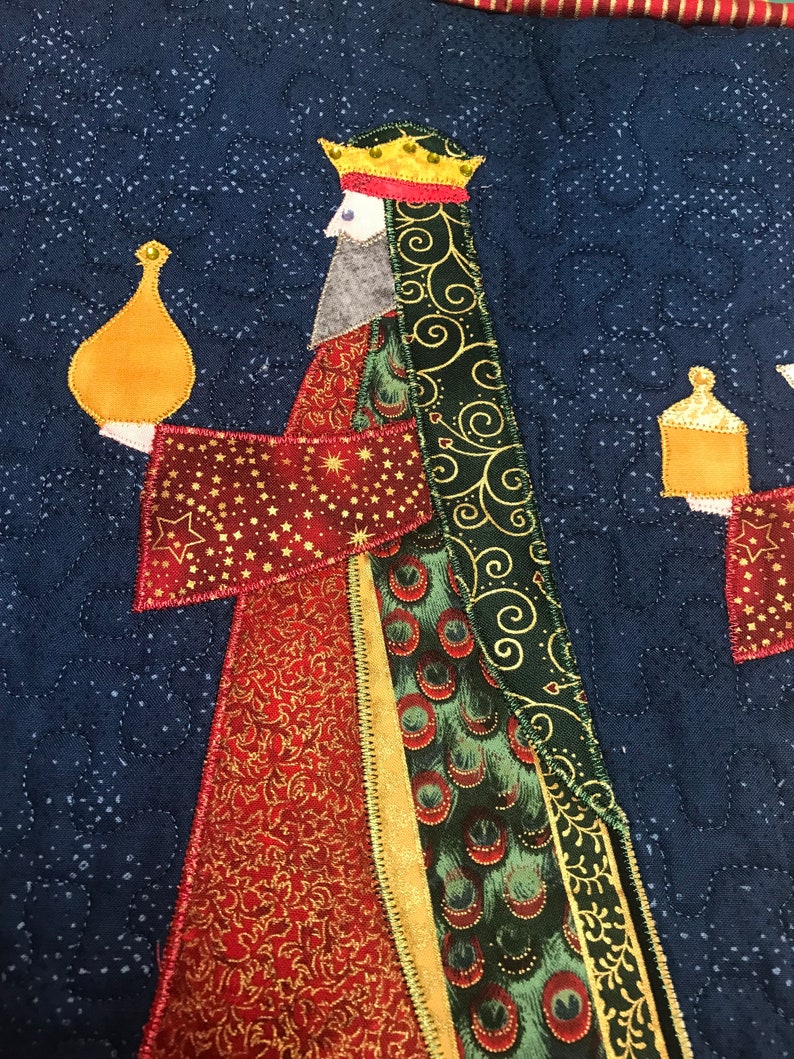 May include: A quilted Christmas stocking featuring a wise man holding a gold gift box. The wise man is wearing a red and gold robe with a green and gold patterned cape. The background is a dark blue fabric with a star pattern.