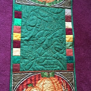 May include: A green quilted table runner with a brown, orange and white pumpkin design. The runner has a border of colourful fabric squares and the text "Autumn Greetings" and "Bountiful Blessings".