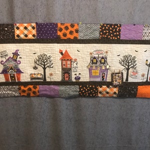 May include: A Halloween-themed quilt runner with a white background featuring a row of houses with black, orange, and purple accents. The houses have signs that read "Witch's Brew", "Candy Corn", and "Boo!" The quilt runner has a brown border and a patchwork border with orange, black, purple, and white fabric.