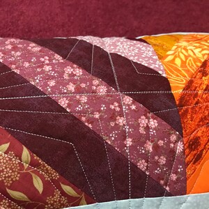 Puede incluir: Un primer plano de una almohada con un dise&ntilde;o de patchwork. La almohada est&aacute; hecha de diferentes telas en tonos rojos, naranjas y marrones. Las telas est&aacute;n cosidas juntas en un patr&oacute;n de chevron. La almohada est&aacute; acolchada con hilo blanco.