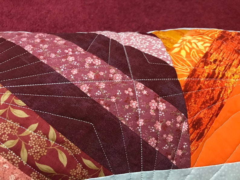Puede incluir: Un primer plano de una almohada con un dise&ntilde;o de patchwork. La almohada est&aacute; hecha de diferentes telas en tonos rojos, naranjas y marrones. Las telas est&aacute;n cosidas juntas en un patr&oacute;n de chevron. La almohada est&aacute; acolchada con hilo blanco.