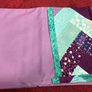 May include: A purple and teal patchwork quilt with a geometric pattern. The quilt is folded in half and has a plain purple side.