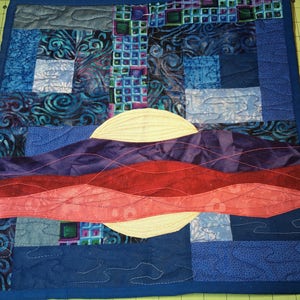 Puede incluir: Un tapiz de pared acolchado cuadrado con un diseño de paisaje. Presenta un sol blanco, olas rojas y moradas en capas y un cielo azul con cuadrados estampados. Los bordes están acabados con un borde azul.