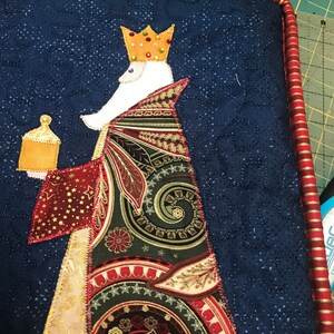 May include: A quilted Christmas stocking with a blue background and a white figure wearing a gold crown and a colorful robe. The figure is holding a gold container.
