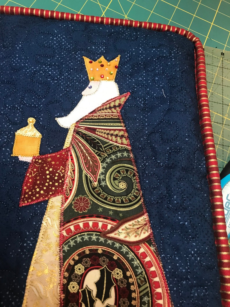 May include: A quilted Christmas stocking with a blue background and a white figure wearing a gold crown and a colorful robe. The figure is holding a gold container.