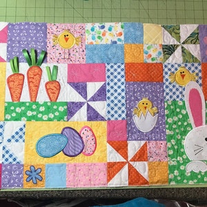 May include: A colorful patchwork quilt with an Easter theme. The quilt features bunnies, chicks, carrots, and eggs. The quilt is made of various fabrics in shades of pink, yellow, green, blue, and purple.