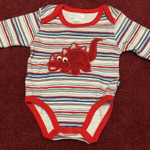 May include: A white and red striped long-sleeved baby onesie with a red dinosaur applique. The onesie has a red trim around the neckline and the snaps are on the bottom. The brand name "Littlefeller" is printed on the tag.
