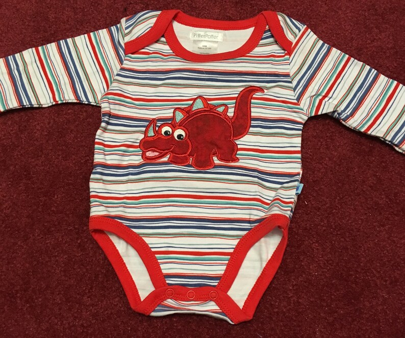 May include: A white and red striped long-sleeved baby onesie with a red dinosaur applique. The onesie has a red trim around the neckline and the snaps are on the bottom. The brand name "Littlefeller" is printed on the tag.