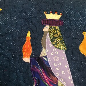 May include: A quilted fabric wall hanging featuring a king with a gold crown and purple robe, holding a candle. The background is a dark blue with a white star pattern.