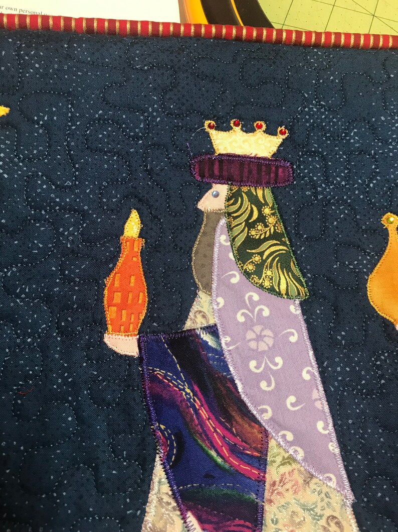 May include: A quilted fabric wall hanging featuring a king with a gold crown and purple robe, holding a candle. The background is a dark blue with a white star pattern.