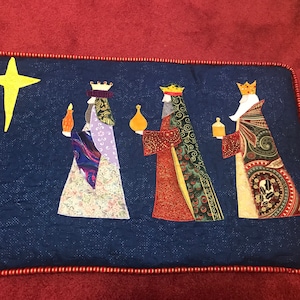 May include: A blue quilted wall hanging with a red border features three wise men in colorful robes carrying gifts. A yellow star shines above them.
