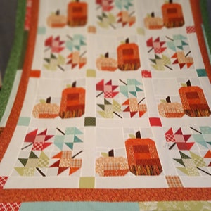 Puede incluir: Una colcha blanca con un diseño de patchwork con calabazas naranjas y hojas de otoño. La colcha tiene un borde verde, azul y naranja.