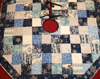 Puede incluir: Una falda de árbol de Navidad acolchada azul y blanca con un diseño de copo de nieve y escena de invierno. La falda tiene un agujero central para el tronco del árbol y está atada con una cinta negra.