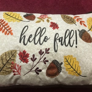Puede incluir: Una almohada beige con hojas de otoño, bellotas y el texto "hello fall!" bordadas en negro.