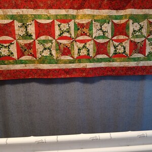 May include: A red, green and white quilted table runner with a floral pattern. The runner is made up of squares with curved edges and features a repeating pattern of flowers and leaves.