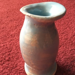 May include: A small, light blue ceramic vase with a unique, asymmetrical shape. The vase has a slightly textured surface and a subtle, iridescent sheen.