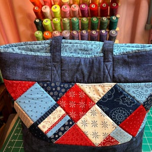 May include: A quilted tote bag featuring a patchwork design of red, navy blue, light blue, and cream fabric squares with floral and geometric patterns. The bag has a solid dark blue top band and handles, sitting on a green cutting mat.