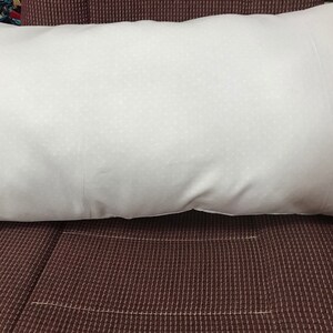 Puede incluir: Una almohada blanca con una superficie texturizada sutil, descansando sobre una tela marr&oacute;n oscura con un patr&oacute;n de cuadr&iacute;cula.