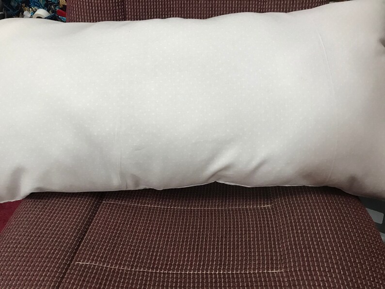 Puede incluir: Una almohada blanca con una superficie texturizada sutil, descansando sobre una tela marr&oacute;n oscura con un patr&oacute;n de cuadr&iacute;cula.