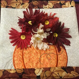 May include: A quilted fabric wall hanging featuring a brown, orange and white pumpkin design. The pumpkin is surrounded by red, white and burgundy fabric flowers.