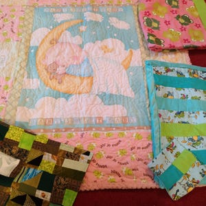 May include: Four baby quilts with different patterns. One quilt features a baby sleeping on a crescent moon with clouds and stars. Another quilt has a green and blue striped pattern with frogs. A third quilt has a pink and green pattern with frogs. The fourth quilt has a patchwork pattern with green, brown, and white squares.