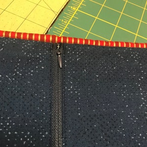 May include: A close-up of a black zipper sewn into a dark blue fabric with a white speckled pattern. The zipper has a silver pull tab.