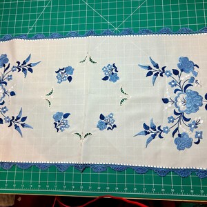 May include: White rectangular table runner with blue floral embroidery and a blue crocheted border. The embroidered flowers and leaves are in varying shades of blue. The runner is on a green surface.