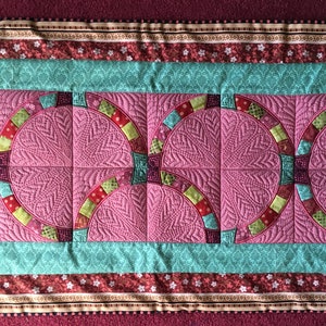 May include: A pink and green quilted table runner with a geometric pattern. The runner has a teal border and a red, white and brown border.