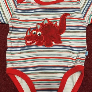 May include: A white and red striped baby onesie with a red dinosaur applique. The onesie has a red trim and snaps at the bottom. The tag on the onesie reads "PitterPatter".