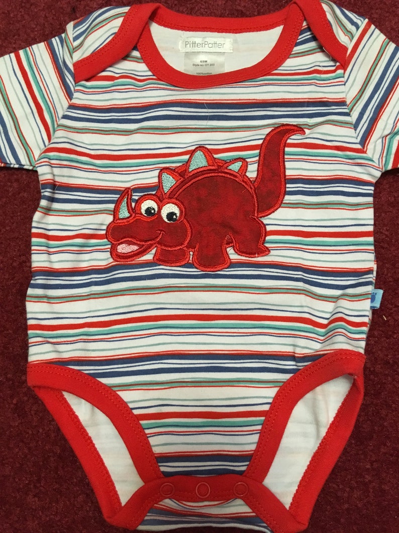 May include: A white and red striped baby onesie with a red dinosaur applique. The onesie has a red trim and snaps at the bottom. The tag on the onesie reads "PitterPatter".