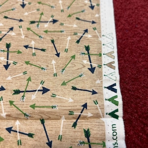 May include: Tan fabric with a repeating pattern of green, white, and blue arrows. The fabric has a white border with a repeating pattern of green, white, and tan triangles. The text "ns.com" is printed on the white border.
