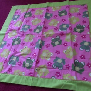 May include: A pink and green baby blanket with a frog and flower pattern. The blanket has a green border.