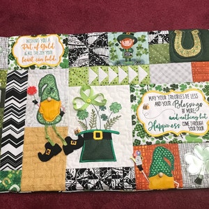 May include: A St. Patrick's Day themed quilted table runner. The runner features a patchwork design with gnomes, shamrocks, rainbows, pots of gold, and the word "LUCKY". The text "Wishing you a pot of gold & all the joy your heart can hold" is also included.