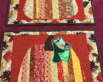 May include: Two quilted placemats with a brown border and a patchwork pumpkin design in shades of orange, red, brown, and yellow. The pumpkins have a green stem and leaves.
