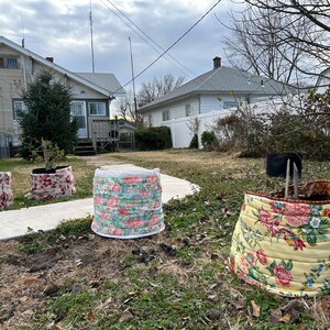 May include: Outdoor garden planters with floral fabric covers. The planters are cylindrical and vary in size, with a mix of red, pink, and yellow floral patterns. The scene is set in a yard with houses in the background.