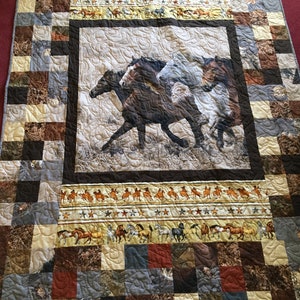 以下が含まれることがあります： 茶色、白、黒の色合いで走る馬の中央の画像が特徴のパッチワークキルト。キルトには、馬とカウボーイの繰り返し模様のボーダーと、さまざまなアーストーンのパッチワークデザインがあります。