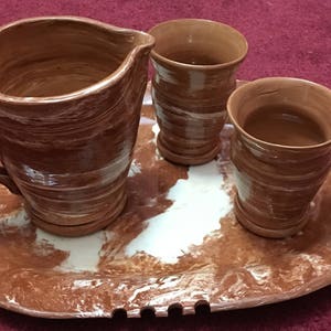Puede incluir: Un juego de tres tazas de cerámica marrón y una jarra con diseños de espiral blanca, todo sobre una bandeja marrón a juego con diseños de espiral blanca.