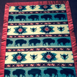 May include: A white, blue, and red patterned fleece blanket with a red satin border. The blanket features a repeating pattern of buffalo silhouettes and geometric designs.