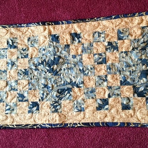 May include: A quilted table runner with a chequered pattern in gold and blue. The fabric features a floral design with gold accents.