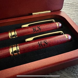 May include: Two burgundy pens with gold trim, engraved with the initials "KS", presented in a wooden box. The pens are displayed in a dark wooden box with a gold hinge. The box is lined with black velvet.