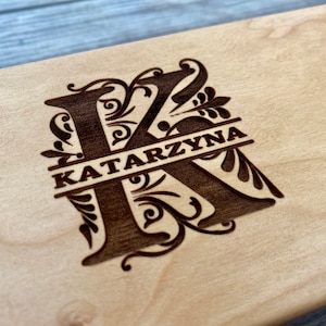 May include: Close-up of a light-coloured wooden surface with an engraved design. The design features a large letter "K" with the name "KATARZYNA" inscribed across it, surrounded by ornate floral elements.