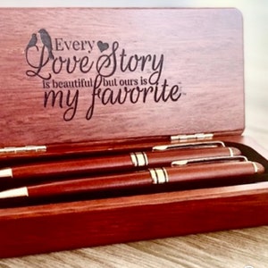 May include: A wooden pen box with a hinged lid, containing two pens. The box is engraved with the words "Every Love Story is beautiful but ours is my favorite" and two bird silhouettes. The pens are dark brown with gold detailing.